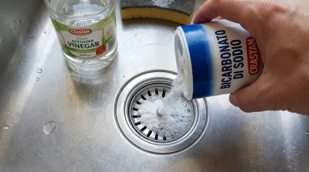 Mano che versa bicarbonato di sodio nello scarico del lavello accanto a una bottiglia di aceto