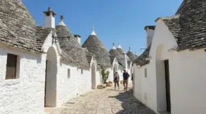 Vicolo acciottolato tra i trulli bianchi di Alberobello, con alcune persone che passeggiano sotto il cielo azzurro